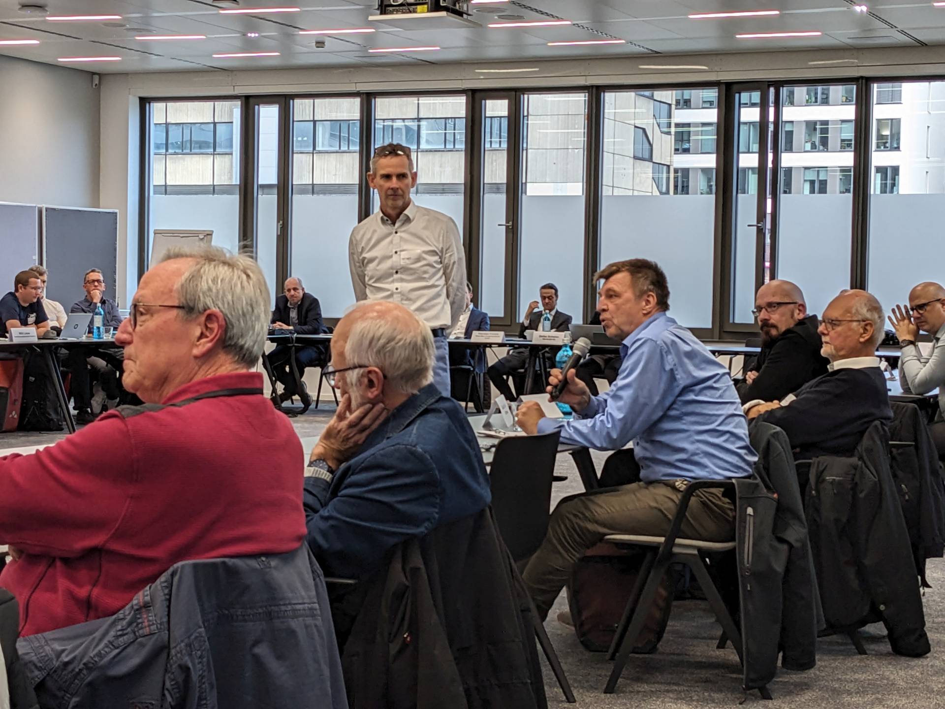 Ein Konferenzraum mit vielen Menschen. Ein Mann in weißem Hemd steht im Zentrum, während ein sitzender Mann in blauem Hemd in ein Mikrophon spricht.