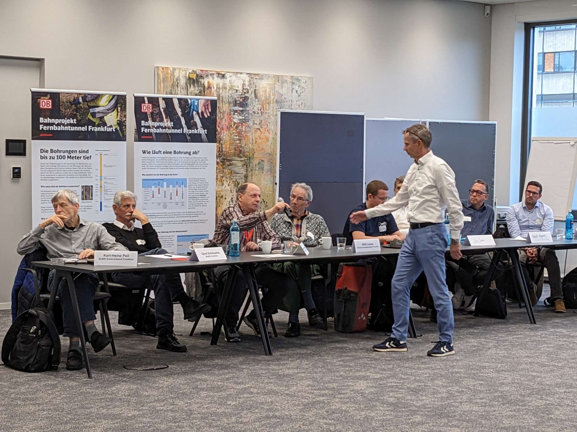 Ein Konferenzraum mit vielen Menschen. Gerade überreicht ein sitzender Mann einem stehenden Mann ein Mikrophon. Im Hintergrund stehen zwei Roll-Ups zum Projekt Fernbahntunnel.
